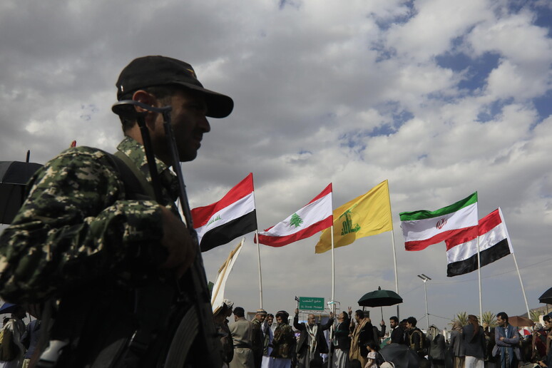 A Houthi soldier stands guard as Houthi supporters wave the flags © ANSA/EPA